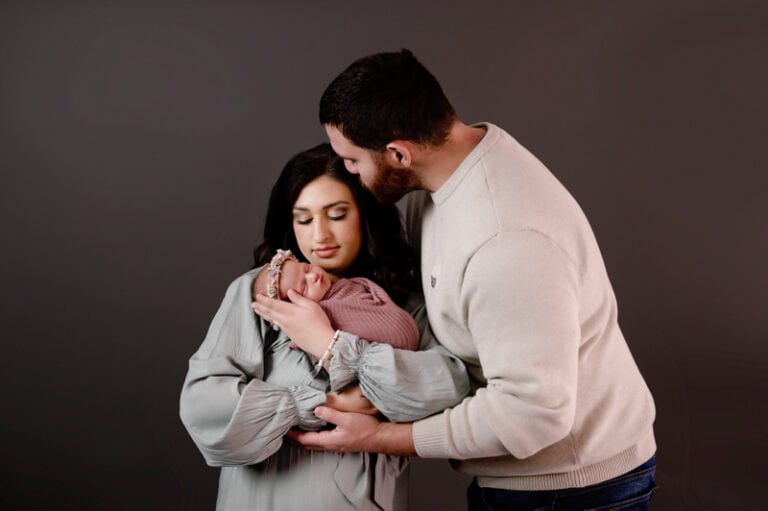 New mom and dad with their new baby girl. Mom is holding the new baby up to her cheek, dad has his arm around mom and baby while kissing mom.