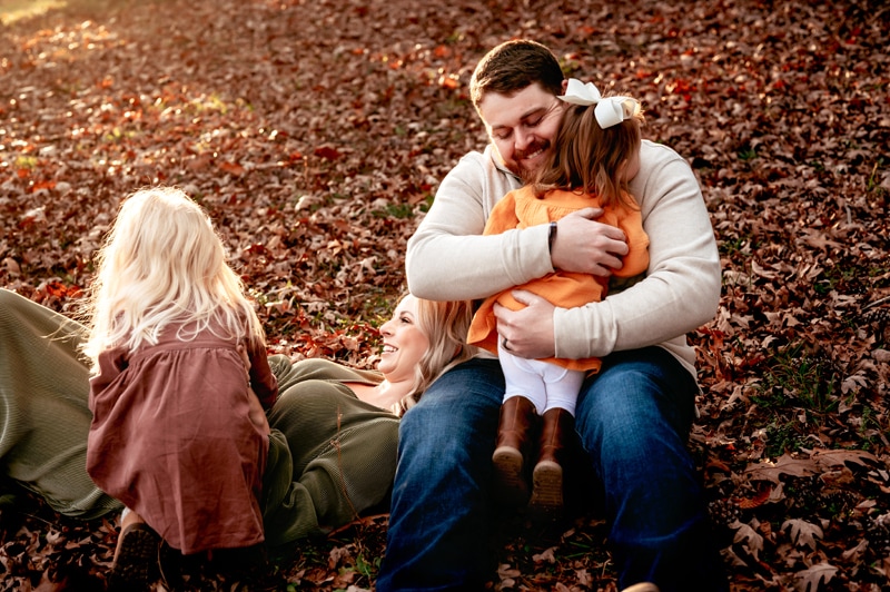 Dad sitting on the ground, mom lying her head on his lap. Oldest daughter is tickling mom to make her laugh and youngest is being hugged by dad. Fall leaves all around them.