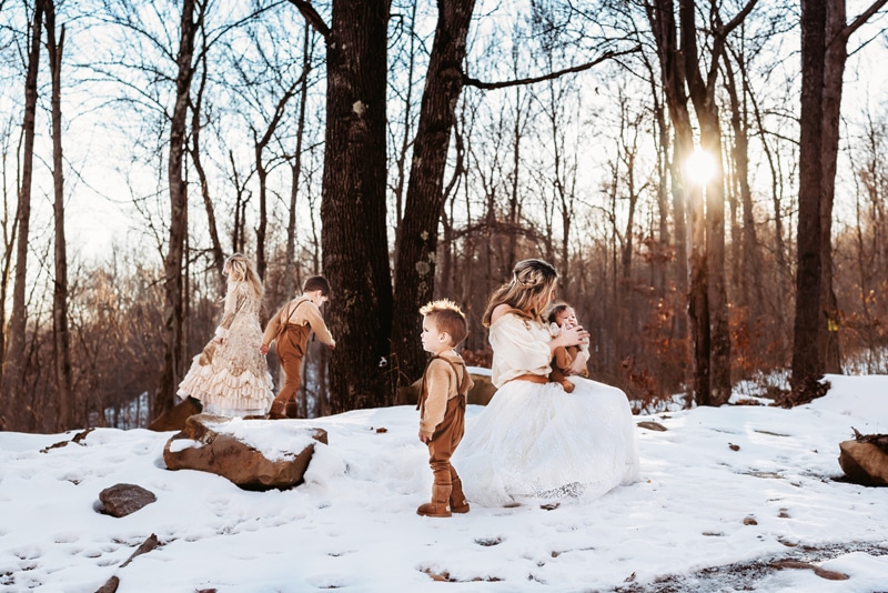 Mom sitting on a rock, wearing all white, surrounded by snow. Her daughter and sons are all around her playing while she is playing with her smallest son on her lap.