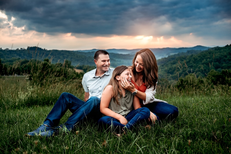 Mom, dad, tween girl sitting on a mountain side. Mom is touching daughter's cheek, all are laughing.