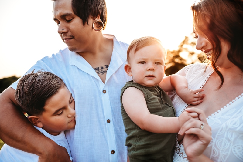 Closeup of a family of 4. Dad has his arm around the oldest boy, a tween, and mom holding their youngest boy, 18 months.