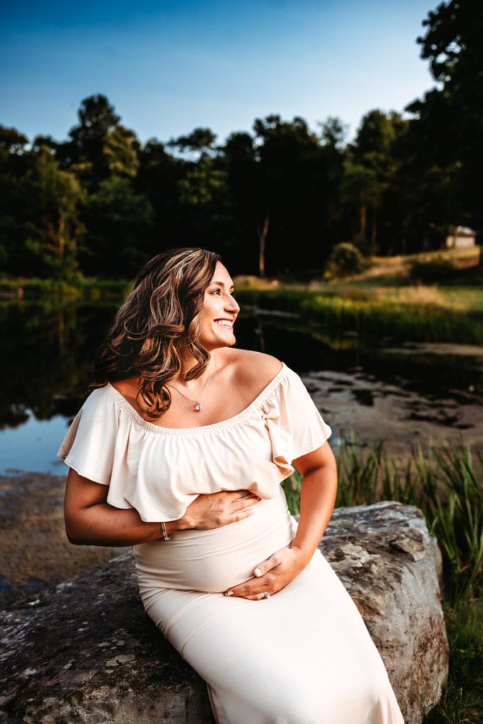 Maternity Photographer, a woman in a white dress is expecting, she holds onto her belly while sitting outdoors near a quiet river