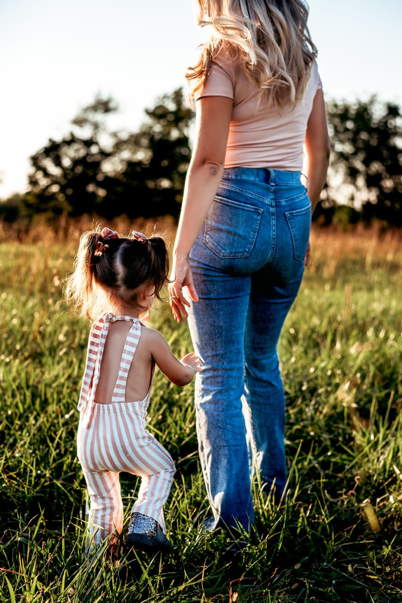 Mom and toddler girl walking through a field of green grass holding hands.