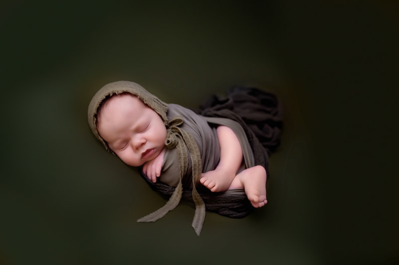 Newborn baby boy wrapped in green, lying on a green backdrop, with a green bonnet.