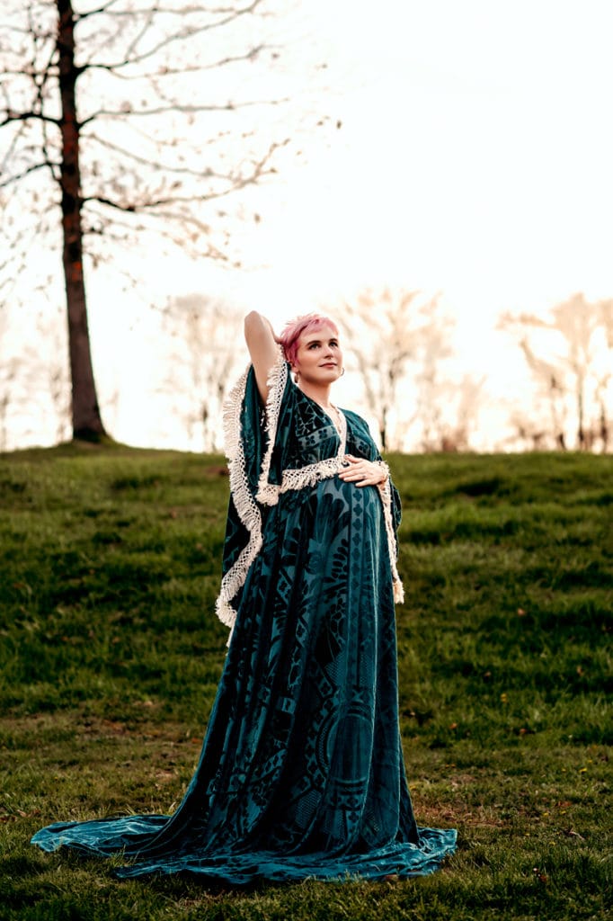 Maternity Session outdoors. Mom is wearing a velvet long boho dress with a long train and cape sleeves. She is holding her bump with one hand, the other hand behind her head, looking off into the distance.