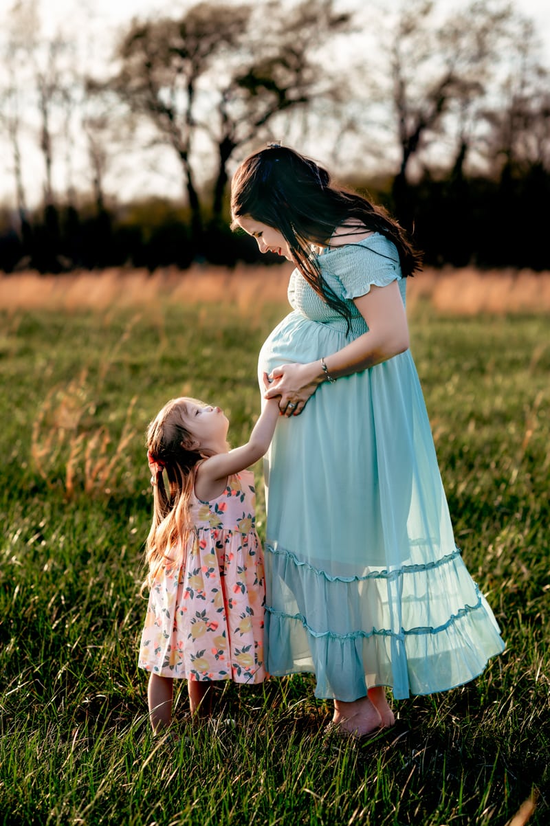 Expectant mom and her toddler daughter in a field with tall grass. Mom is wearing a mint green dress and daughter in a floral dress. Daughter is going for a kiss on mom's belly while mom looks down at with a smile.