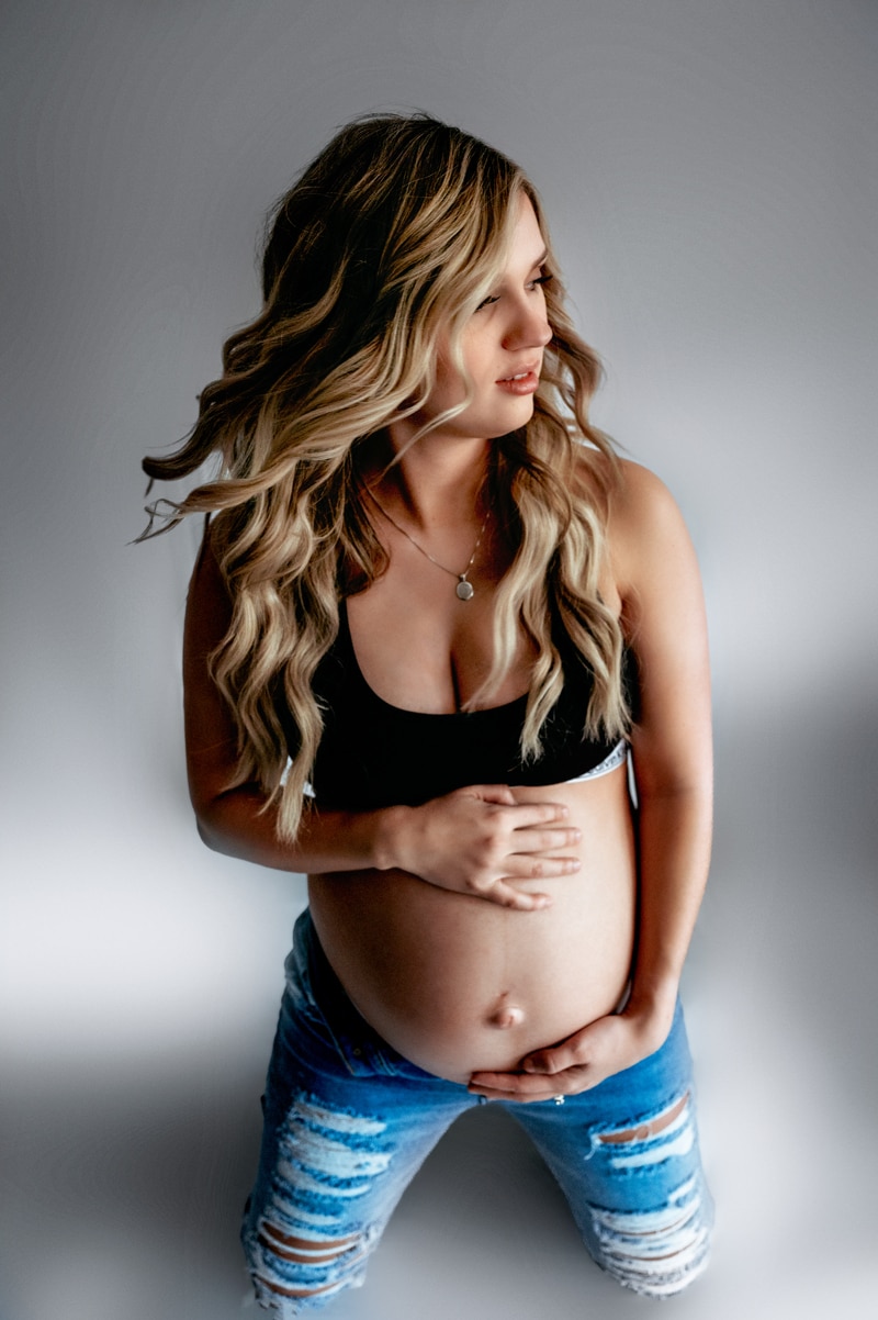 Studio maternity session. Gray background with mom in ripped jeans and a calvin klein sports bra. Mom is holding her baby bump and tossing her hair.