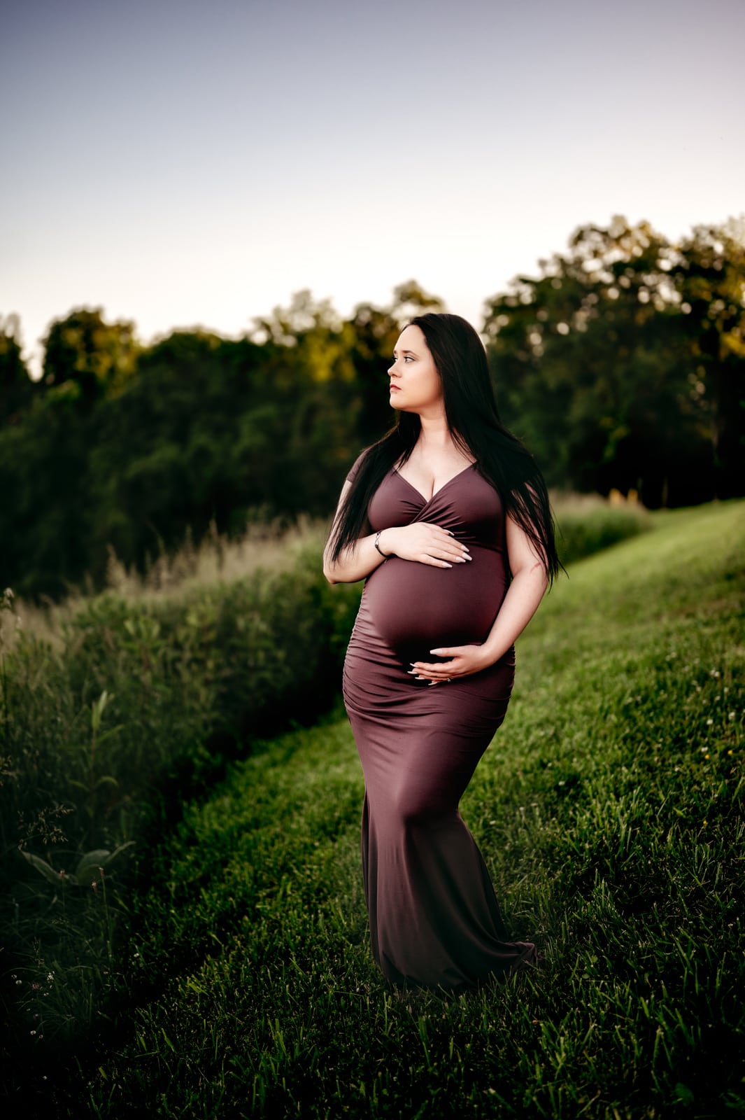 Maternity outdoors, Mom is wearing a fitted purple dress and holding her baby bump with green trees and grass all around her, looking off in the distance.