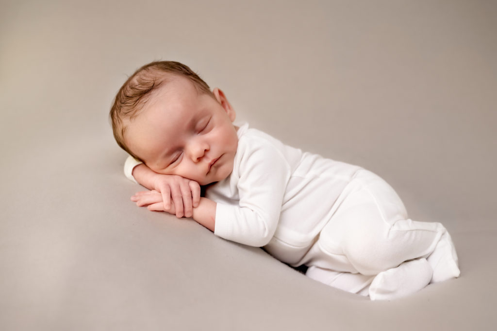 Newborn baby boy lying on his side, wearing a white footed pajama on a cream background.