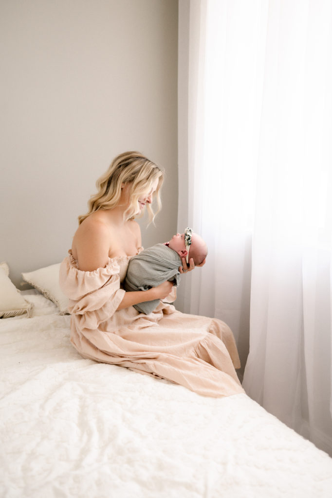 Mom wearing a cream colored dress, sitting on a white bed and holding her newborn girl. Baby girl is wrapped in a sage green swaddle wrap and wearing a flower crown.