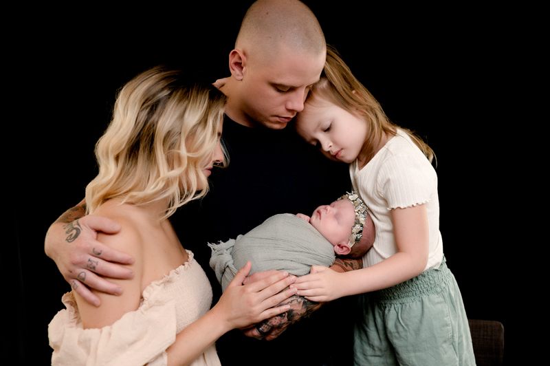 Mom, dad, toddler girl, and newborn girl with a black background behind them. The family is looking down at the new baby, all with their hands holding her.