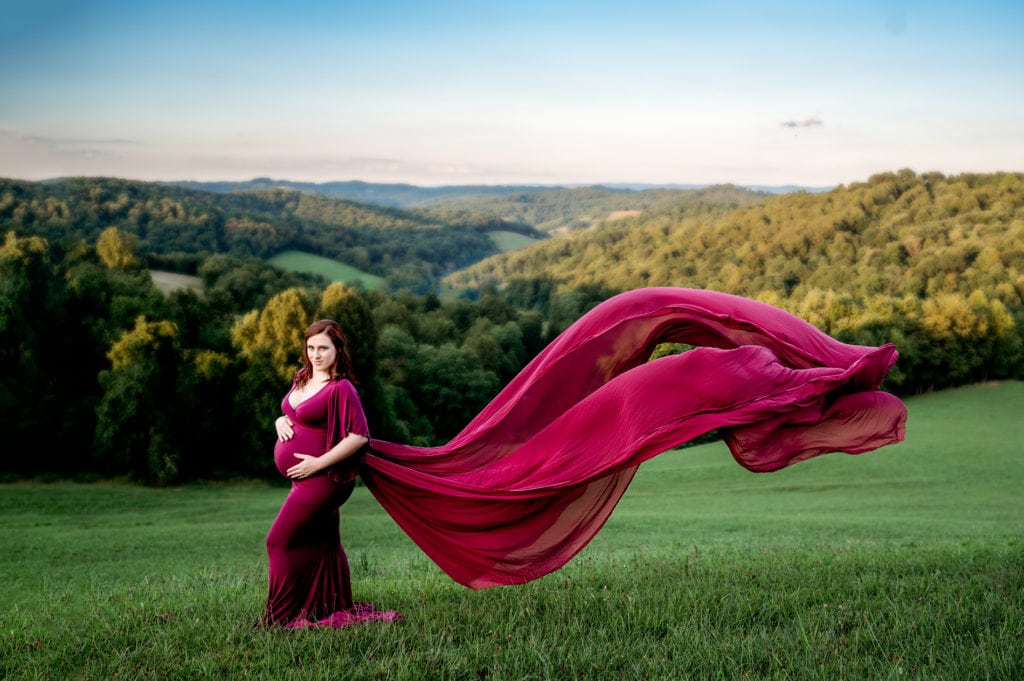 Maternity session with mom holding her bump, standing on a hill with mountains behind her. The train of her dress is flowing in the air.