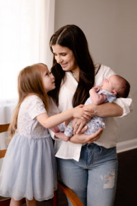 Mom is standing and holding her newborn baby girl. Her older daughter is standing on a chair with one hand on the baby, head on mom and looking up at mom.