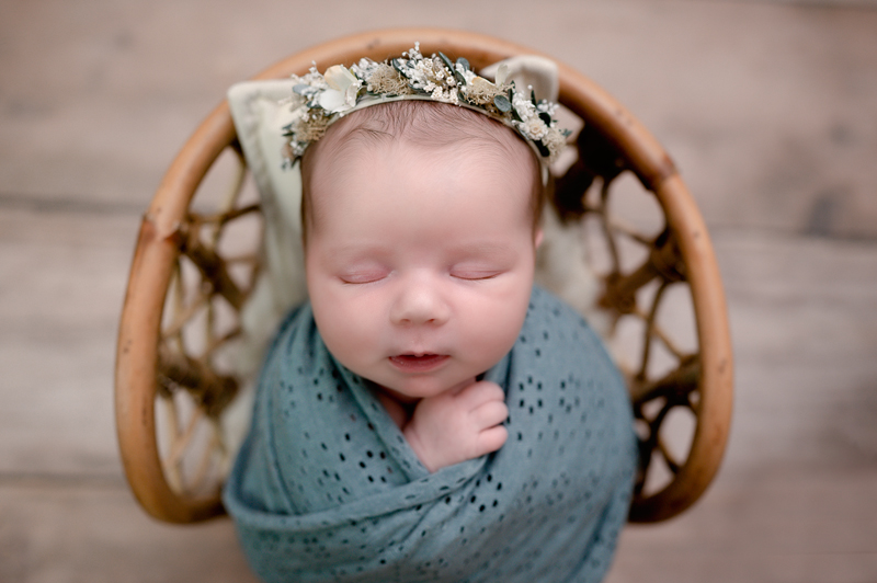 Newborn baby girl swaddled in a blue wrap wearing a flower crown and sitting in a chair.