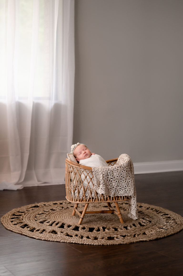 Newborn baby girl swaddled in a white wrap inside of a basket, in front of a white curtain on a jute rug.