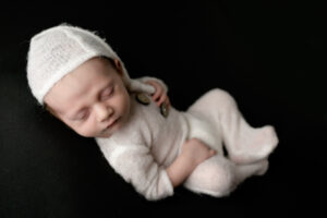 Newborn baby boy in a white outfit on a black background with a white sleepy cap.