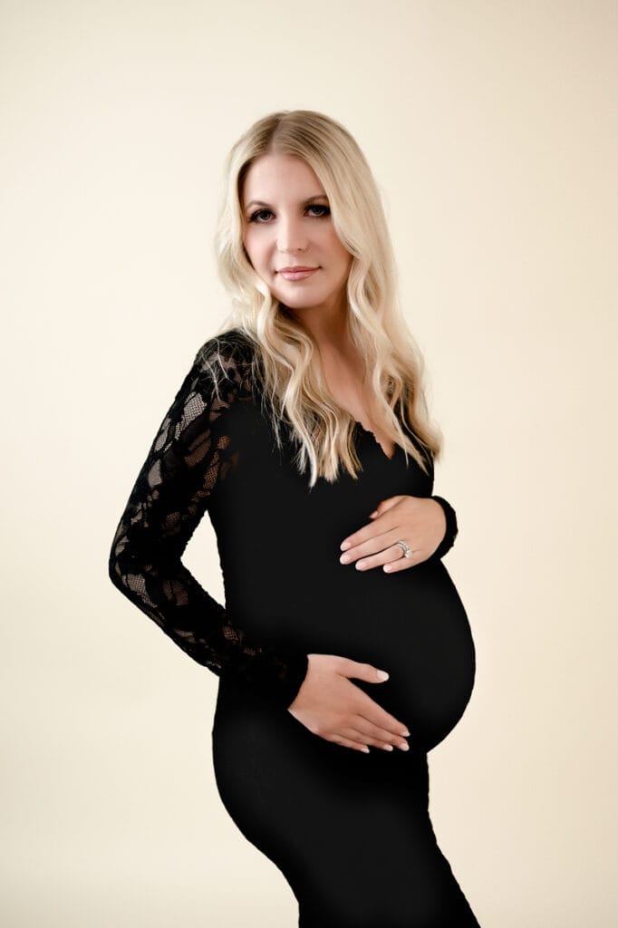 Maternity session in studio. Mom to be is wearing a black dress and holding her bump, looking at the camera.