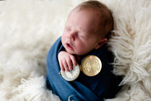 Newborn baby boy holding his great pap's compass. Baby is swaddled in a blue wrap on a white fluffy background.