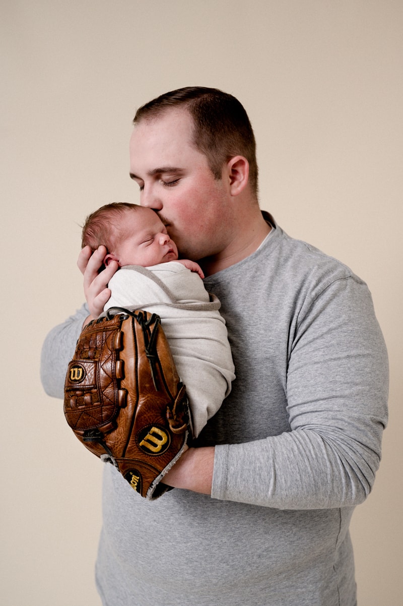 Dad is holding his newborn baby boy in his old high school baseball glove and giving him a kiss.