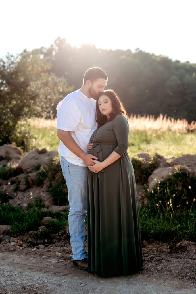 Expectant mom is wearing a green maxi dress with her husband kissing her. There is green all around them and they are both holding the baby bump.