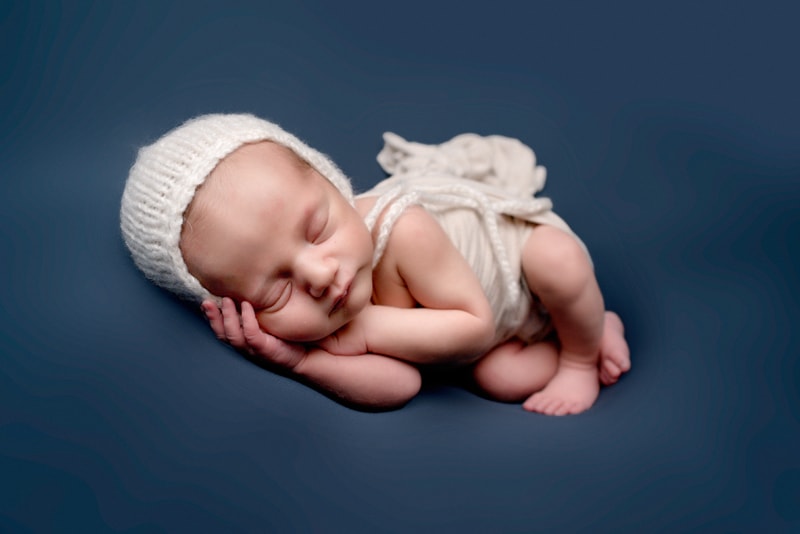 Newborn baby boy lying on a blue backdrop with a white bonnet.