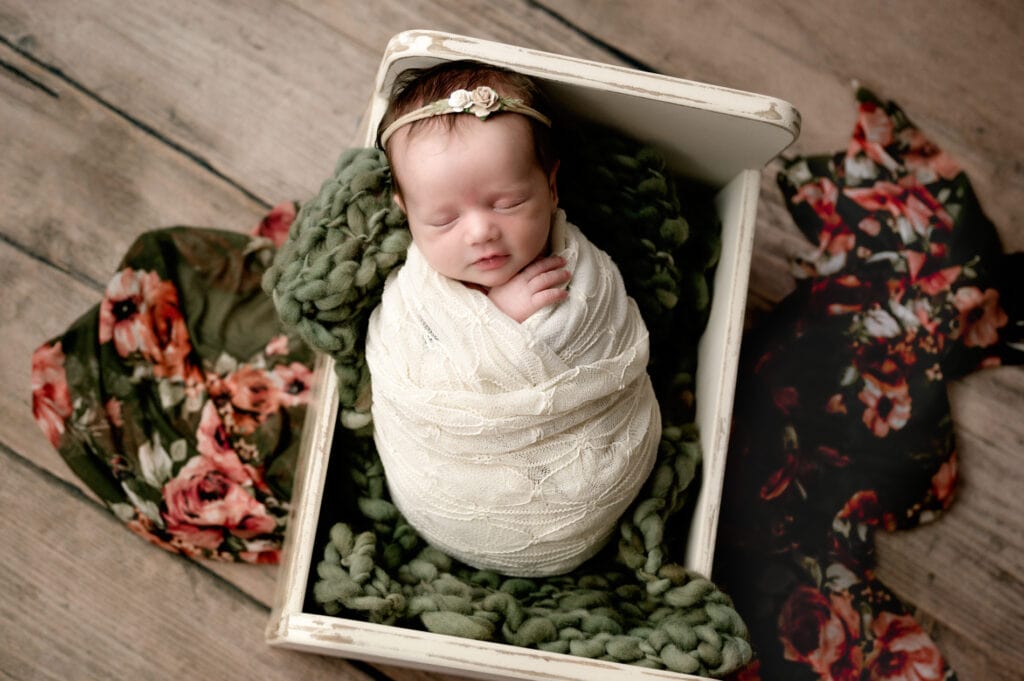 Newborn baby girl in a cream crate with a green fluffy under her. She is wrapped in a cream lace wrap and a flower layer is under the crate.