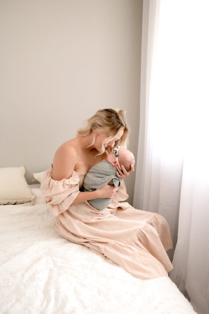Mom is sitting on a white bed, wearing a cream colored dress. She is holding and kissing her newborn girl that is swaddled in a sage green wrap and wearing a flower crown.
