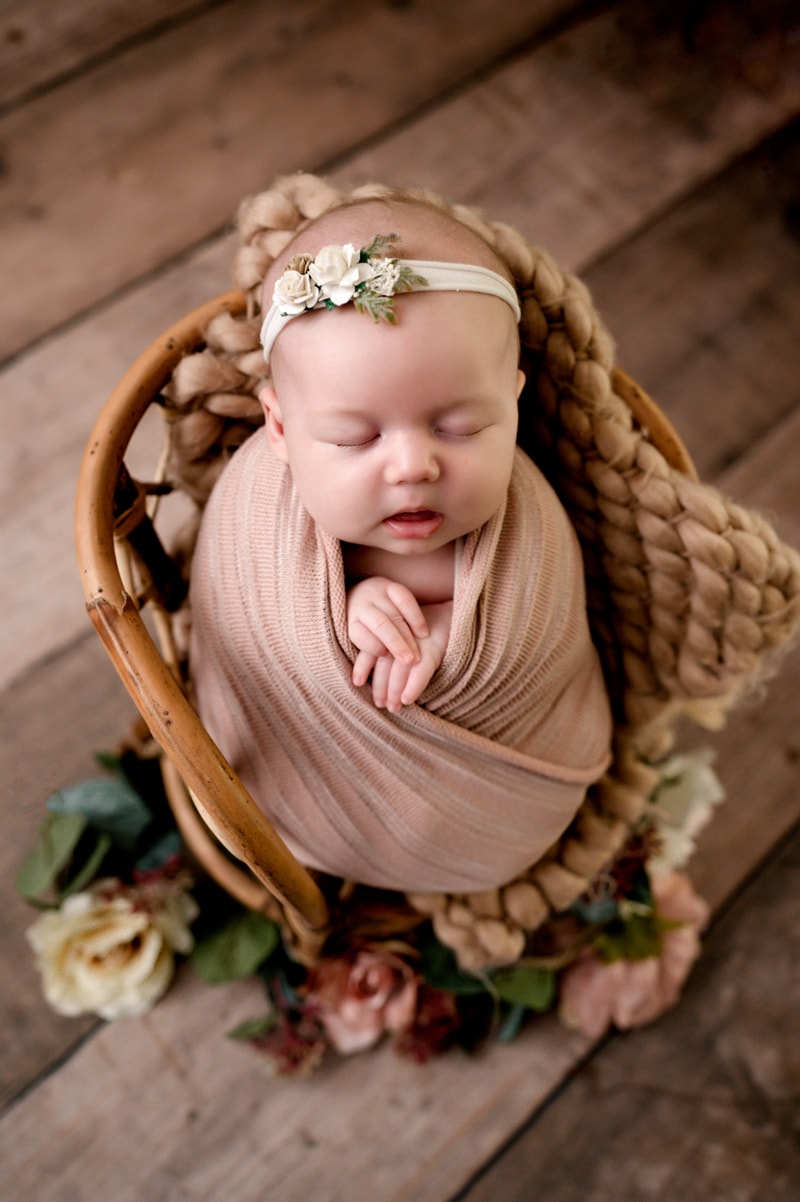 Newborn girl in a rattan chair with flowers and a flower headband. She is swaddled in a pink wrap.