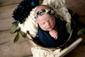 Newborn baby girl wrapped in navy blur with a blue crown. She has greenery and flowers around her.