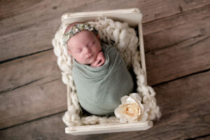 Newborn baby girl in a neutral toned setup. She is wearing a sage green wrap with matching flower crown and lying on a cream background