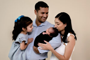 Family session in studio. Dad is holding toddler girl and newborn baby boy while mom kisses baby boy.