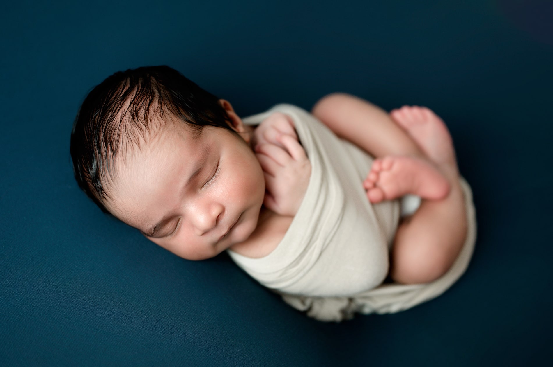 Newborn boy on a blue backdrop swaddled in a blue wrap with his feet out.