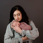 Newborn session in studio with mom holding her newborn girl. Mom is wearing a green dress, baby is wrapped in a mauve wrap with a gray background.