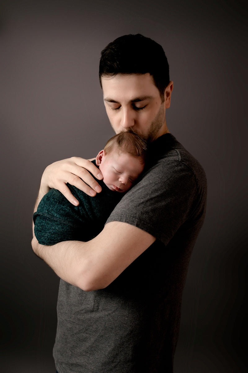 Newborn session in studio. Dad is holding is newborn baby boy on his chest and kissing his head. Baby boy is wrapped in a green wrap. They are in front of a gray background.