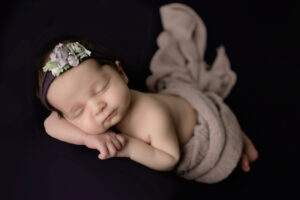 Newborn girl in studio with a flower headband and lying on a purple posing table.