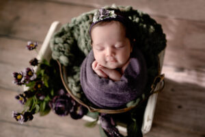 Newborn baby girl in studio. She is wrapped in a purple wrap and has greenery and purple flowers around her.