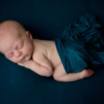 Newborn boy posed on a teal background. He is on his stomach with his hand under his cheek.