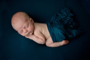 Newborn boy posed on a teal background. He is on his stomach with his hand under his cheek.