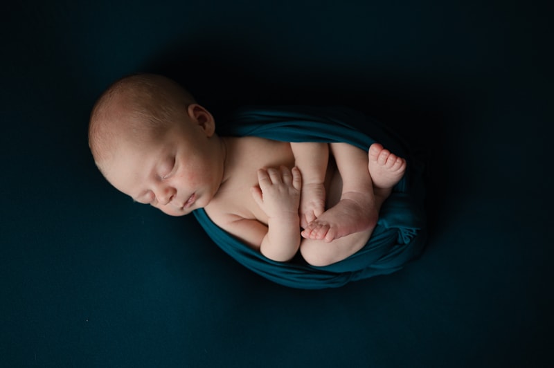 Newborn Boy in the studio, posed on teal. He is lying on his back, with his feet tucked up and his arms showing.