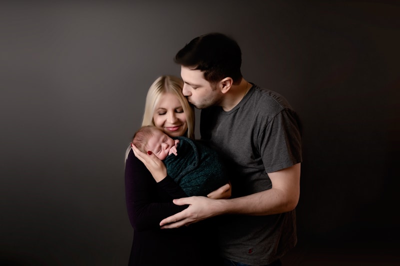 Mom, dad, and baby in the studio on a dark gray background. Mom is holding baby, dad is kissing mom.