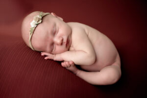 Newborn baby girl posed on red. She is in a taco pose on her stomach with her hands and feet under her.