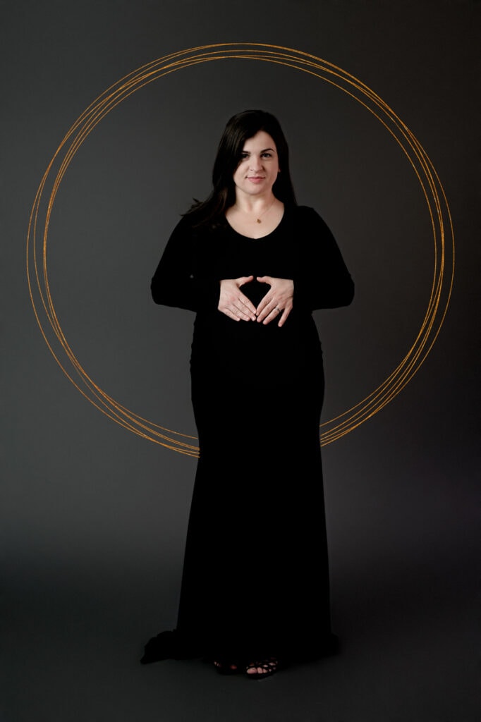 Maternity photoshoot in studio. Mom is wearing a black dress with a gray background and a gold ring around her.