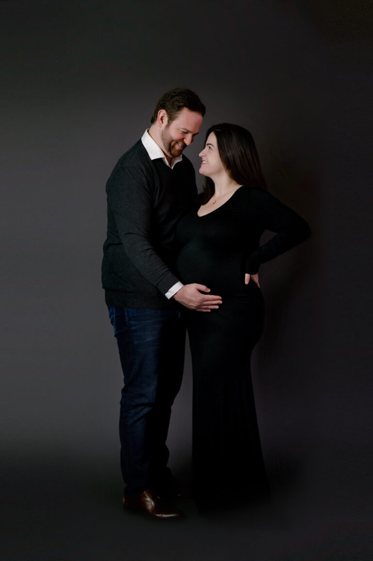 Maternity photoshoot in studio. Mom is wearing a black long gown and dad is holding her baby bump. They are looking into each other's eyes.