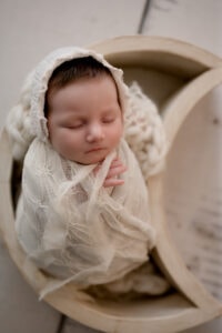 Newborn girl wrapped in a cream lace wrap with a cream lace bonnet. She is posed inside of a cream moon bowl prop with a cream wood floor behind her.
