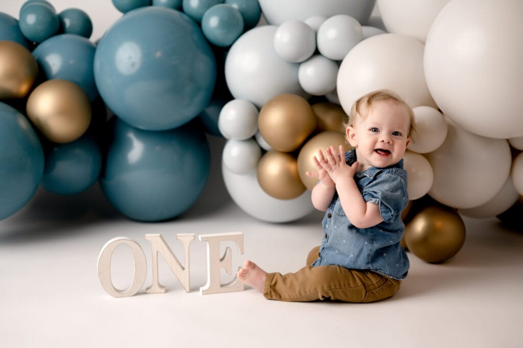 Morgantown, WV cake smash photographer with a boy in studio. He is sitting on the floor with blue and gold balloons behind him from Wildflower Events, letters spelling out ONE and he is clapping is hands while smiling.