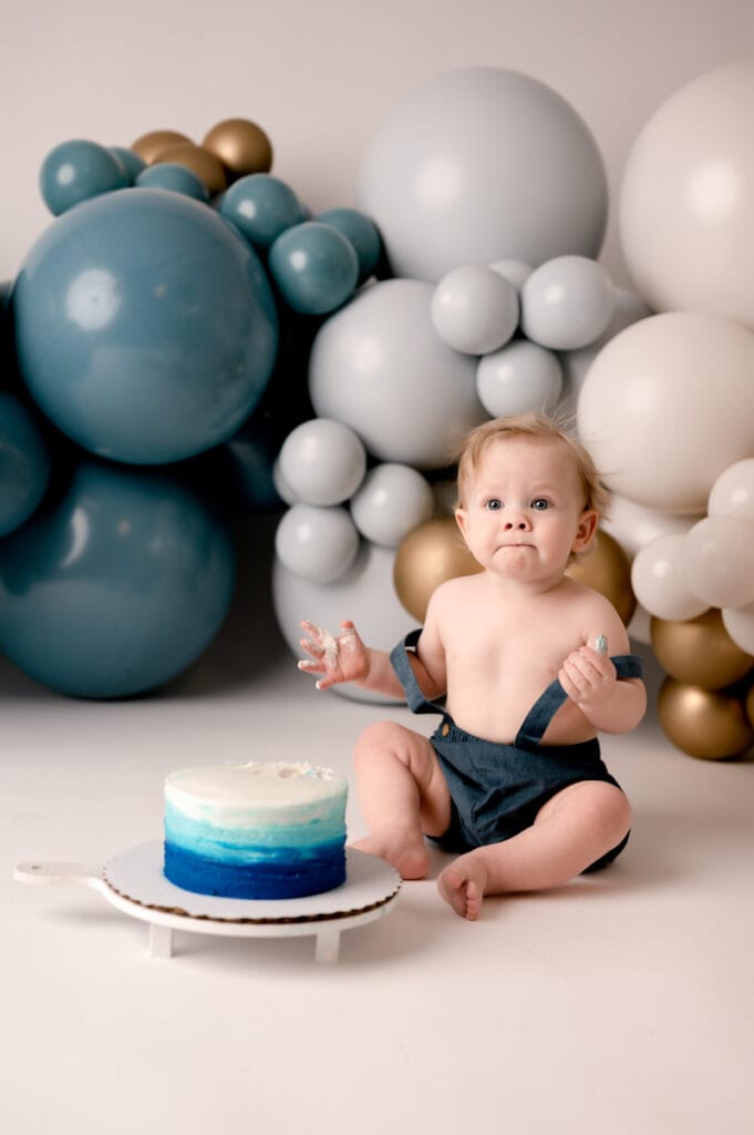 One year old cake smash session in the studio in Morgantown, WV. He is wearing bubble short suspenders and eating cake. The background is white with dark blue, light blue, white, and gold balloons from Wildflower Events.