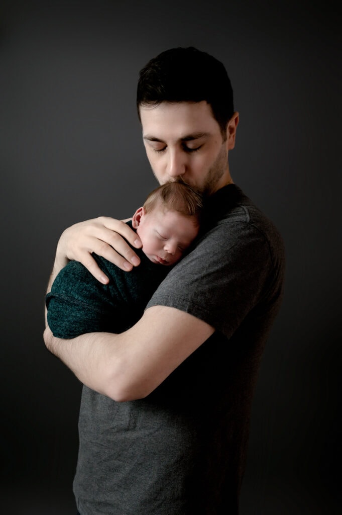Newborn session in studio. Dad is holding is newborn baby boy on his chest and kissing his head. Baby boy is wrapped in a green wrap. They are in front of a gray background.