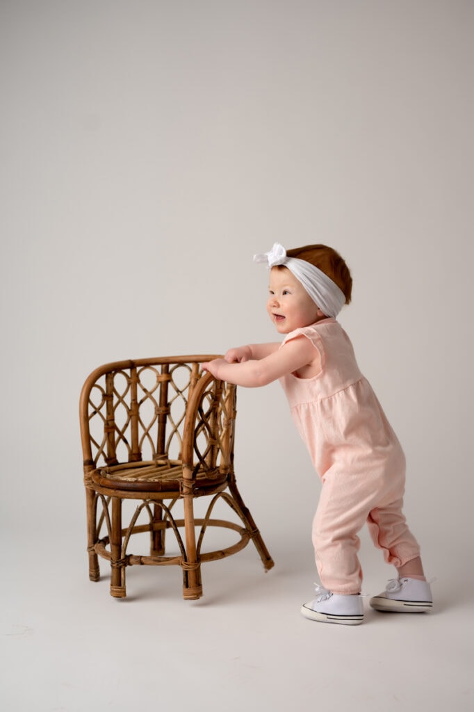 One year girl in the Morgantown, WV photography studio. She is wearing a peach outfit, white headband, and standing with a rattan chair on a white background.