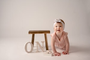 Little girl turning one in the studio. There are letters spelling out one on the floor with a wooden stool behind it. She is crawling towards the camera with a smile.