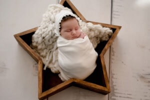 Newborn baby girl in the studio. She is posed inside a wooden star prop, wrapped in all white with a white bonnet.